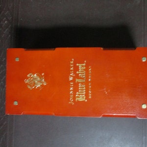 Pu&ograve; includere: Una scatola rettangolare arancione con scritte dorate e un'emblema dorato. Il testo sulla scatola recita "JOHNNIE WALKER Blue Label SCOTCH WHISKY". La scatola ha quattro piccoli borchie metalliche color oro.