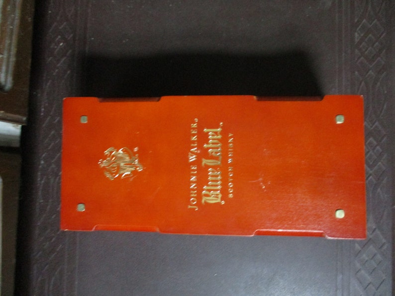 Pu&ograve; includere: Una scatola rettangolare arancione con scritte dorate e un'emblema dorato. Il testo sulla scatola recita "JOHNNIE WALKER Blue Label SCOTCH WHISKY". La scatola ha quattro piccoli borchie metalliche color oro.