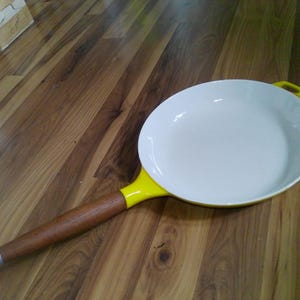 May include: A white frying pan with a yellow handle and a wooden handle. The pan is on a wooden surface. The pan has a diameter of approximately 10 inches.