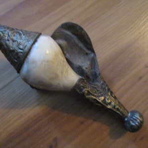 May include: An antique horn-shaped object with ornate metal detailing. The horn is cream-colored, and the metal has a tarnished, antique appearance. The object has a spoon-like extension and a decorative metal tip.