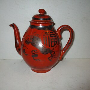 May include: A red ceramic teapot with a black handle, spout, and lid. The teapot features a black and silver design with Chinese characters and a dragon motif. The teapot is a traditional Asian design.