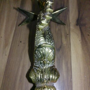 May include: A polished brass door knocker featuring a stylized seahorse design with a textured body and a smooth, shell-like base. Behind the seahorse, a four-pointed star emblem is visible. A wooden ruler is positioned at the top for scale.