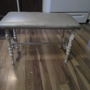 May include: A white painted metal vanity stool with a padded gray seat. The stool has a decorative, ornate design on the legs.