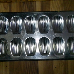 May include: A rectangular, silver-colored madeleine pan with twelve shell-shaped molds. The pan is made of metal and has a slightly reflective surface. The molds have vertical ridges, and the pan sits on a wooden surface.