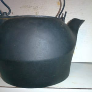 May include: A black cast iron kettle with a hinged lid and a spout. The kettle has a handle on the top and a smaller handle on the side.