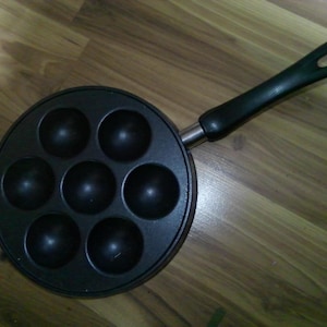 May include: A black, round, multi-sectioned pan with a long, black handle. The pan has seven circular indentations for cooking. The handle is attached to the pan with a silver metal piece. The pan is on a wooden surface.