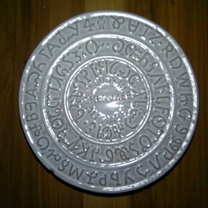 May include: A circular light gray ceramic plate featuring embossed Cherokee syllabary characters arranged in concentric circles. The center of the plate displays the text CHEROKEE ALPHABET in raised lettering, surrounded by various symbols and characters.