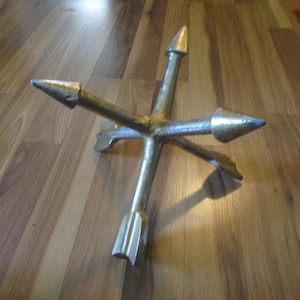 May include: A gold-colored metal decorative object with four arrow-shaped points extending from a central hub. The arrows have pointed tips with a slight blue hue. The object is lying on a wooden surface.