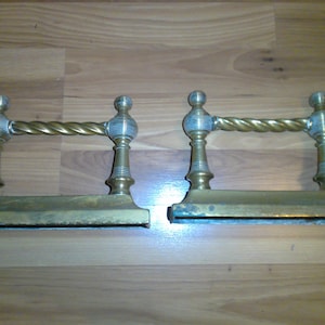 May include: A pair of antique brass fireplace andirons with a twisted design. The andirons have a decorative, fluted design on the top and bottom.
