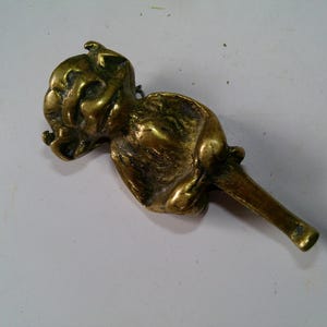May include: A brass sculpture of a grotesque figure with a long, cylindrical stem. The sculpture has a detailed face and body, with a weathered, antique appearance. The object is likely a decorative item.