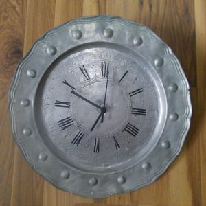 May include: A round, silver-colored wall clock with Roman numerals and black hands. The clock face has a textured design and is set in a scalloped, circular frame with decorative studs. The clock is displayed against a wooden background.