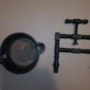 May include: A dark, circular cast iron mortar with two side handles and a matching dark cast iron press tool featuring a central threaded screw mechanism with a T-shaped handle and two vertical side supports with decorative turned detailing.