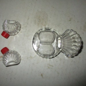 May include: Clear glass serving dish with three compartments and a shell-shaped handle. Two small, shell-shaped glass bottles with red lids are also present. The items are arranged on a light-colored surface.