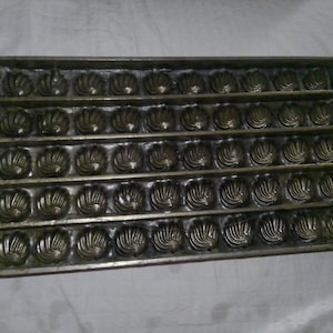 May include: A rectangular metal chocolate mold featuring five horizontal rows, each containing ten individual shell-shaped cavities. The mold has a dark, metallic finish with a uniform grid structure designed for creating multiple small confections.