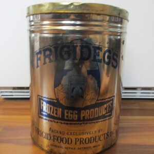 May include: A vintage metal can with a gold lid and a silver body. The can features the text "FRIGIDEGS" in blue, along with "FROZEN EGG PRODUCTS" and "FRIGID FOOD PRODUCTS INC." in blue. The can is cylindrical and shows signs of age.