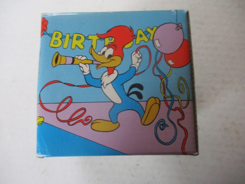 Puede incluir: Un p&aacute;jaro carpintero de dibujos animados azul y rojo con un sombrero rojo sopla un cuerno de fiesta. El fondo es azul con una franja rosa y morada. Las palabras "Birthday" est&aacute;n escritas en letras amarillas. Hay serpentinas rojas, amarillas y azules y globos morados y rojos.