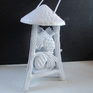 May include: A white metal lantern with a handle, a conical top, and a decorative pinecone design. The lantern has a tree-like frame and is suitable for outdoor or indoor use.