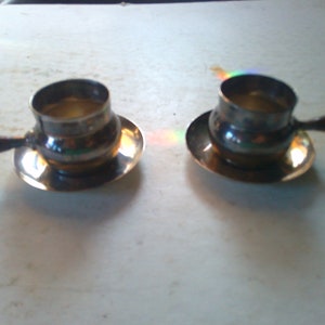 May include: Two silver cups with wooden handles and matching saucers. The cups are filled with a clear liquid.