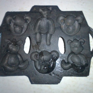 May include: A black cast iron mold for making bear-shaped treats. The mold has six cavities, each shaped like a bear. The mold has two handles on the sides.