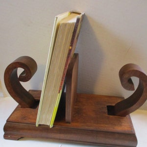 May include: A wooden bookend with a decorative scroll design. The bookend is made of dark brown wood and has a rectangular base. A book is resting in the bookend.