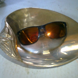 May include: Sunglasses with black frames and orange lenses rest inside a decorative, gold-colored seashell-shaped dish. The dish has a textured surface and a detailed edge design, reflecting light.