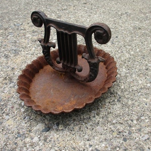 May include: A rusty, decorative metal lyre-shaped object, possibly a candle holder or decorative piece, sits atop a matching oval dish with a scalloped edge. The metal has a weathered, aged appearance, suggesting it is an antique.