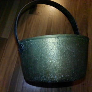 May include: A round, metallic pot with a textured, weathered gold-toned finish and a dark, arched handle attached to the sides. The vessel features a wide, open top and a slightly tapered body, showing signs of age and oxidation on its surface.