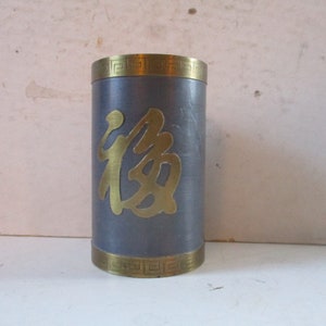 May include: A gray metal cylinder with a gold Chinese character on the front and gold trim around the top and bottom. The character is "福" which means "good fortune" or "happiness".