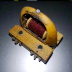 May include: A wooden spool with a red thread wrapped around it. The spool is attached to a wooden base with a handle.