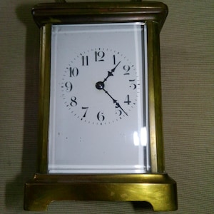 May include: A rectangular brass carriage clock with a white face and black Roman numerals. The clock has a handle at the top and sits on a rectangular base. The clock face is protected by a glass panel.