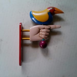 May include: A wooden hand pointing to the right with a yellow, blue, and red bird perched on top. The hand is attached to a red wooden base.
