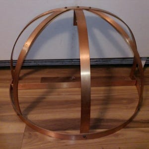 May include: A copper-colored, dome-shaped metal frame. The frame is constructed from curved and straight metal strips, joined at the top and base. The structure is set against a light-colored wall and wooden floor.
