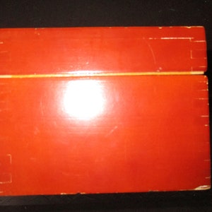 Pu&ograve; includere: Una scatola rettangolare in legno con finitura rosso-arancio lucida. La scatola &egrave; costruita con giunzioni visibili e ha un coperchio incernierato. La parte superiore della scatola &egrave; leggermente pi&ugrave; chiara dei lati.