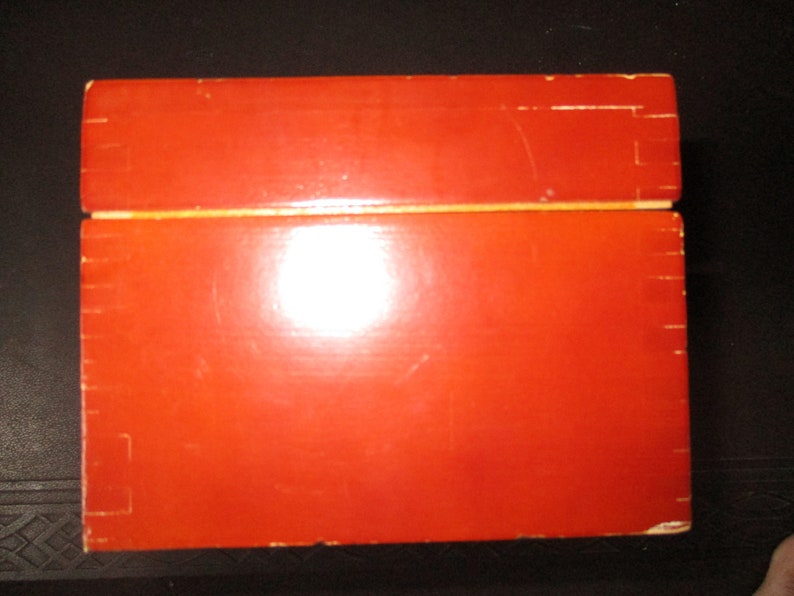 Pu&ograve; includere: Una scatola rettangolare in legno con finitura rosso-arancio lucida. La scatola &egrave; costruita con giunzioni visibili e ha un coperchio incernierato. La parte superiore della scatola &egrave; leggermente pi&ugrave; chiara dei lati.