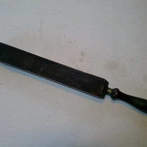 May include: A vintage straight razor strop with a dark wooden handle and a black leather surface. The strop is rectangular and tapers slightly towards the handle. The handle is dark green and oval-shaped.