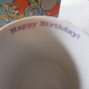Puede incluir: Una taza de cer&aacute;mica blanca con las palabras "Happy Birthday!" impresas en morado en el borde.