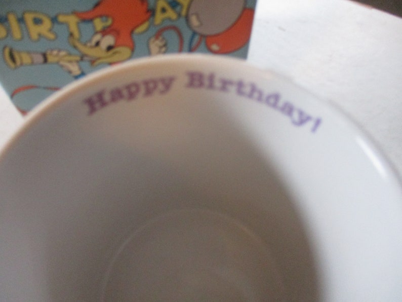 Puede incluir: Una taza de cer&aacute;mica blanca con las palabras "Happy Birthday!" impresas en morado en el borde.