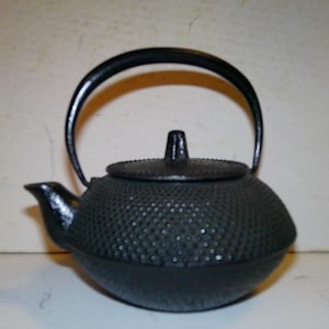 May include: Black cast iron teapot with a textured surface and a rounded body. The teapot has a handle and a spout.