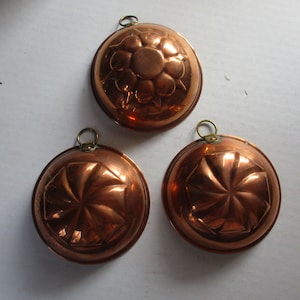 May include: Three copper molds with decorative floral and geometric patterns. Each mold has a circular shape with a small loop for hanging. The molds are made of shiny copper and are likely used for baking or decorative purposes.