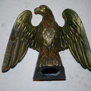 May include: A brass eagle figurine with an aged patina finish, depicted with wings fully spread and feathers detailed in relief. The base features a hollow, oval-shaped opening, and the eagle is oriented with its head turned toward the left side.