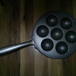 May include: A dark gray, round pan with seven circular indentations for cooking. The pan has a long, silver handle with a loop at the end. The pan is designed for making small, round pancakes or pastries.