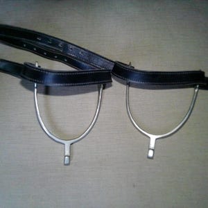 May include: A pair of equestrian spurs featuring silver-toned metal U-shaped bows with short shanks, attached to black leather straps with silver-colored metal buckles and multiple adjustment holes for securing the equipment to footwear.
