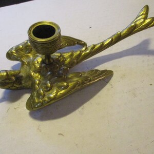 K&ouml;nnte beinhalten: Ein messingfarbener Vogel-Kerzenhalter. Der Vogel wird im Flug dargestellt, mit ausgebreiteten Fl&uuml;geln und einem detaillierten Federdesign. Der Kerzenhalter ist eine kleine, runde Tasse, die f&uuml;r eine einzelne Kerze geeignet ist.