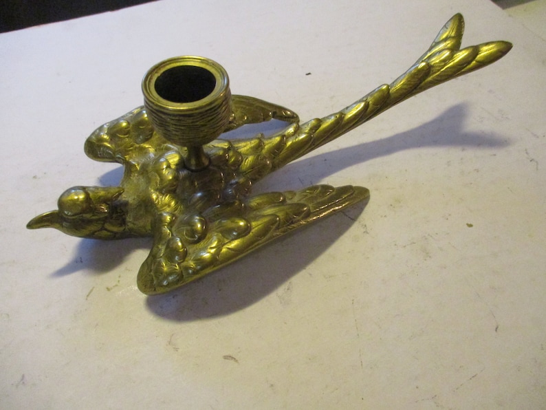 K&ouml;nnte beinhalten: Ein messingfarbener Vogel-Kerzenhalter. Der Vogel wird im Flug dargestellt, mit ausgebreiteten Fl&uuml;geln und einem detaillierten Federdesign. Der Kerzenhalter ist eine kleine, runde Tasse, die f&uuml;r eine einzelne Kerze geeignet ist.