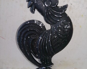 May include: A black rooster statue with a distressed finish. The sculpture features a detailed comb, wattle, and tail feathers. The rooster stands on a round base. The statue is likely made of metal or a similar material.