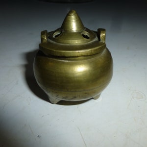 May include: A small, antique brass incense burner with a rounded body and a conical lid. The lid has several small holes and a pointed top. The burner has three small feet and two handles. The overall color is a warm, aged brass.