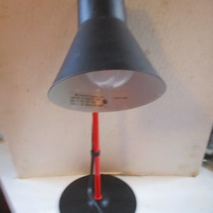 May include: A black and red desk lamp with a black shade and a red metal stem. The lamp has a black base and a black cord.