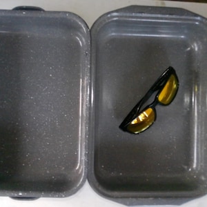 May include: Two gray ceramic baking dishes with a speckled finish. One dish is empty, and the other contains a pair of black sunglasses with gold-tinted lenses.