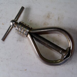 May include: A silver-toned metal nutcracker featuring a teardrop-shaped frame with a central threaded screw mechanism. The tool includes a T-shaped handle at the top for manual operation, designed to apply pressure to crack open hard-shelled nuts.