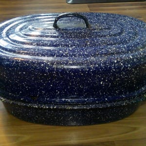 May include: A blue speckled oval roasting pan with a lid and a handle. The pan is sitting on a wooden surface. The roasting pan is a kitchenware item.